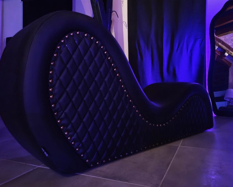Sofa tantra noir en forme ondulée avec des clous décoratifs, mirroir rétro-éclairé par une lumière violette.